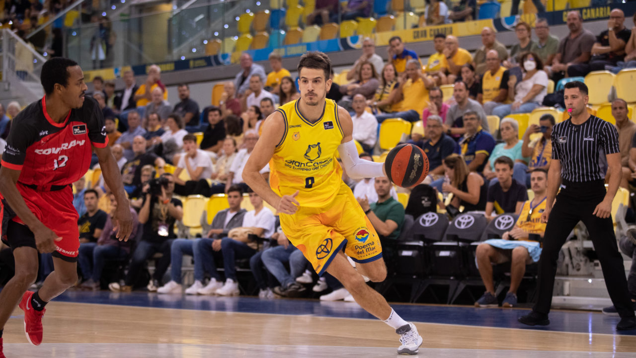 Jugadores de baloncesto compiten en la cancha, con espectadores animando en el fondo.
