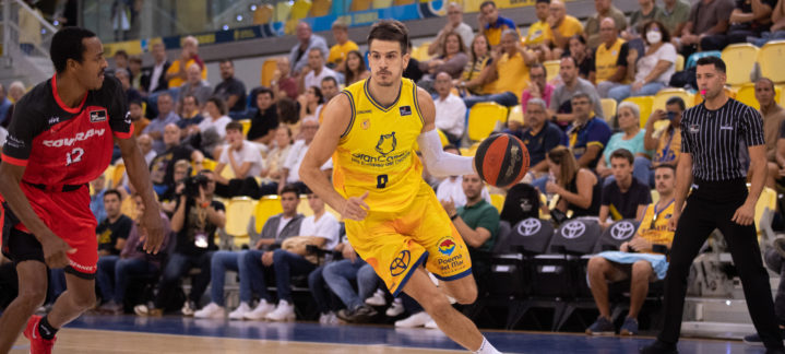 Jugadores de baloncesto compiten en la cancha, con espectadores animando en el fondo.