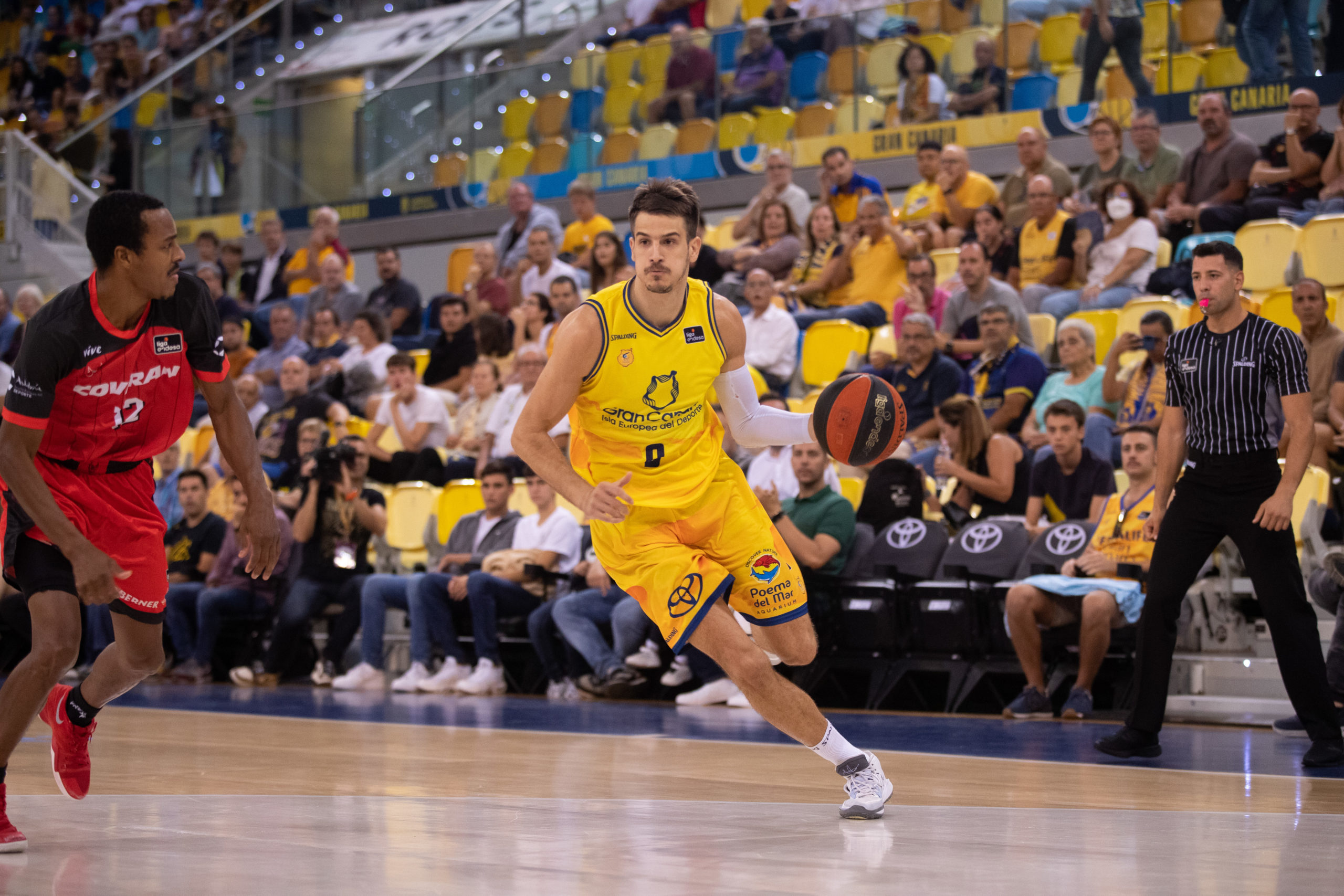 Jugadores de baloncesto compiten en la cancha, con espectadores animando en el fondo.