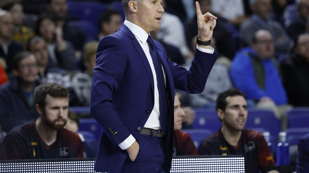 Entrenador gesticulando durante un partido, con público en el fondo.