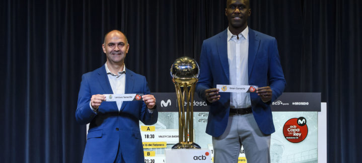 Dos hombres sostienen carteles con los nombres de los equipos, junto a un trofeo de baloncesto.