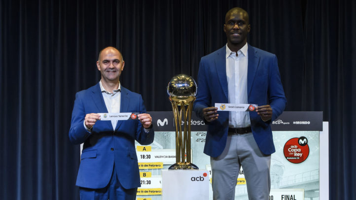 Dos hombres sostienen carteles con los nombres de los equipos, junto a un trofeo de baloncesto.