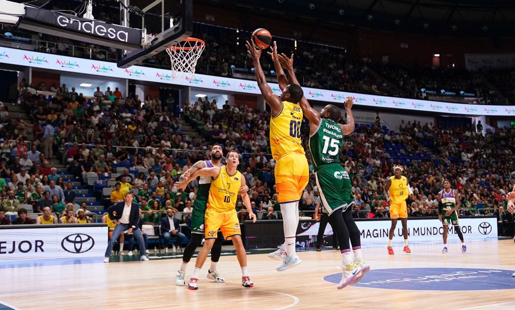 Dos jugadores intentan anotar mientras otros observan en un partido de baloncesto.
