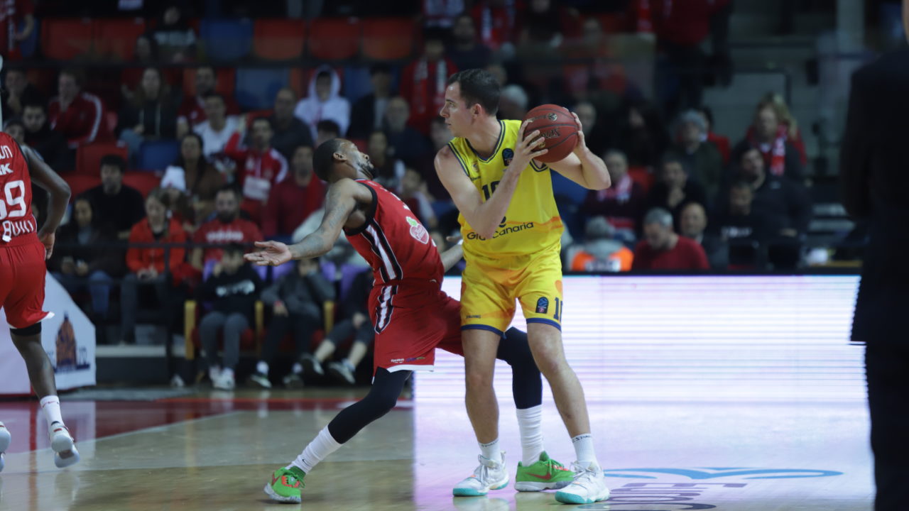 Jugador con balón en acción durante un partido de baloncesto, defendido por otro jugador.