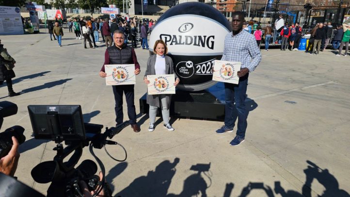 Tres personas sostienen carteles frente a una gran pelota de baloncesto en un evento.