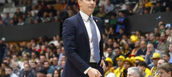Entrenador camina por la línea de banda durante un partido, con público al fondo.