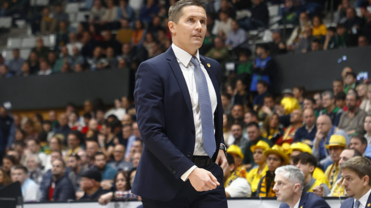 Entrenador camina por la línea de banda durante un partido, con público al fondo.