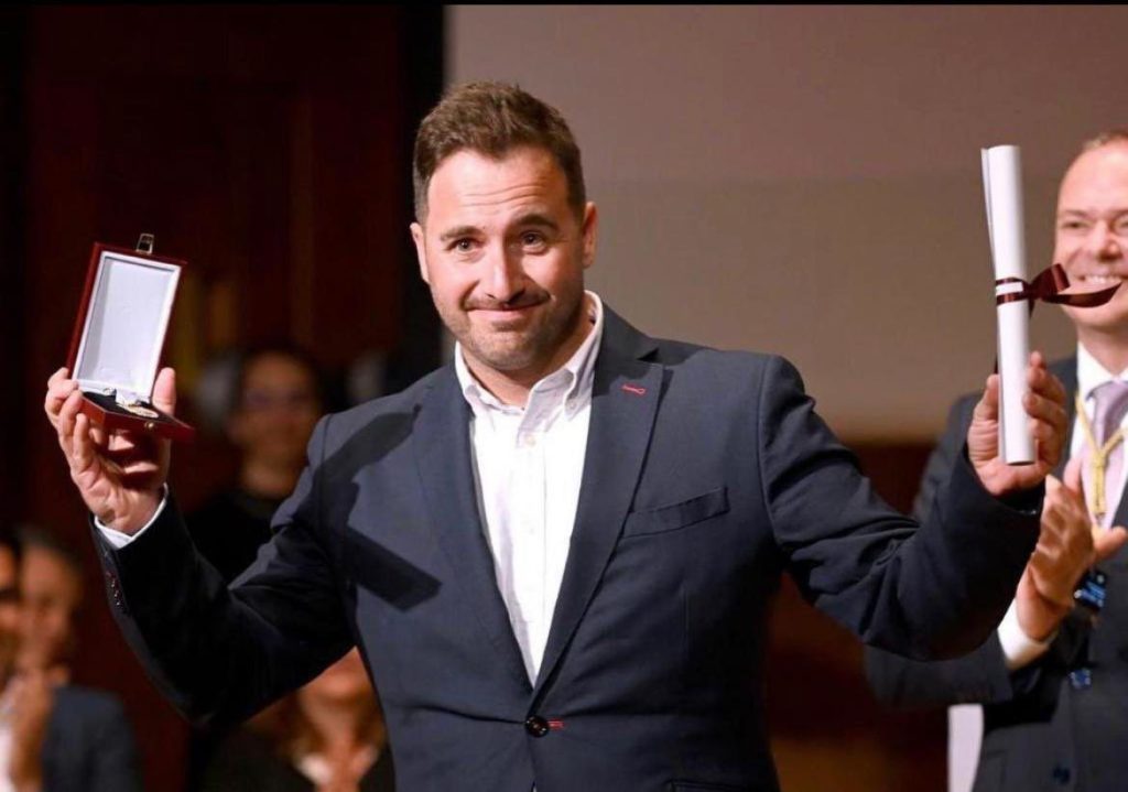 Hombre sonriente sostiene una medalla y un diploma en un evento de premiación.