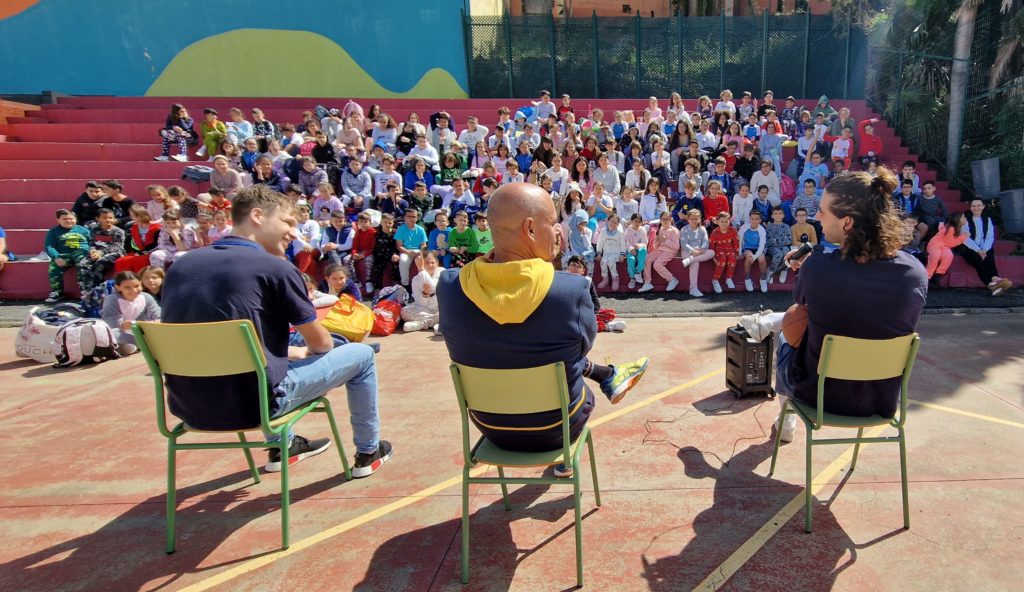 Grupo numeroso de niños y adultos escuchando a tres oradores en un colegio.