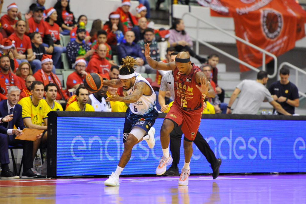 Dos jugadores de baloncesto luchan por el balón en un partido frente a un público animado.