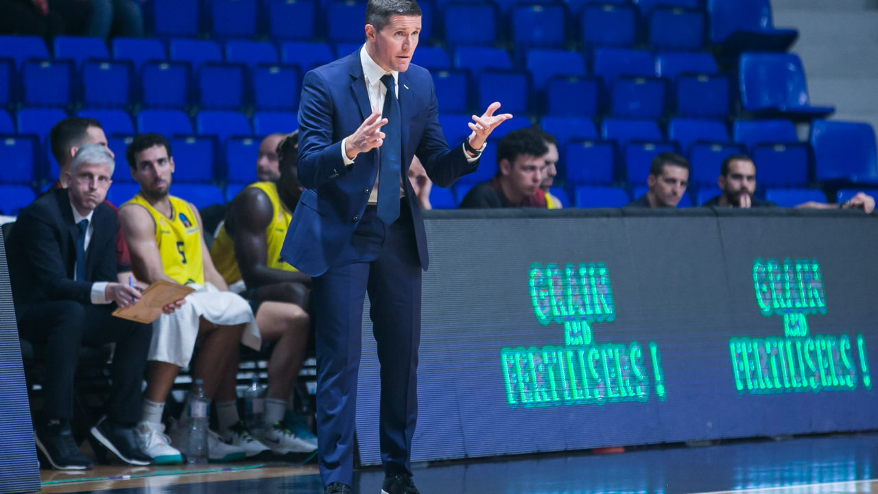 Entrenador gesticulando durante un partido, con jugadores en el banquillo.