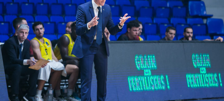 Entrenador gesticulando durante un partido, con jugadores en el banquillo.