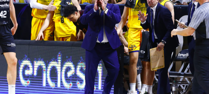 Entrenador gesticulando en un partido, rodeado de jugadores y asistentes del equipo.