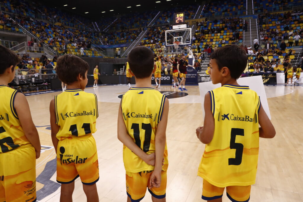 Niños en chándal amarillo del CB Gran Canaria observan un partido en la cancha.