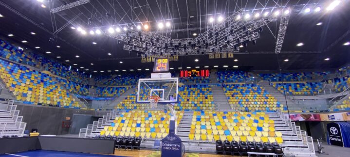 Vista del interior del pabellón con asientos vacíos y canasta de baloncesto.