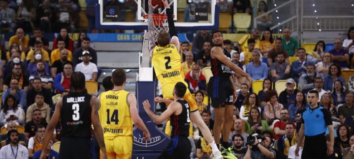 Jugador del Gran Canaria realiza un mate mientras un contrario intenta bloquearlo, con público de fondo.