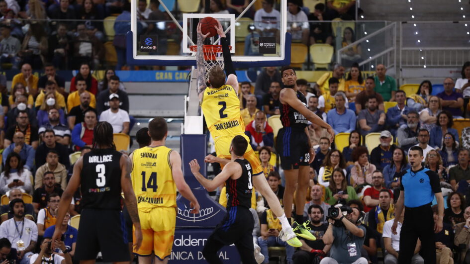 Jugador del Gran Canaria realiza un mate mientras un contrario intenta bloquearlo, con público de fondo.