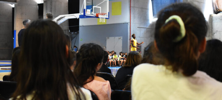Niños y niñas atentos a la charla de un entrenador en una cancha de baloncesto.