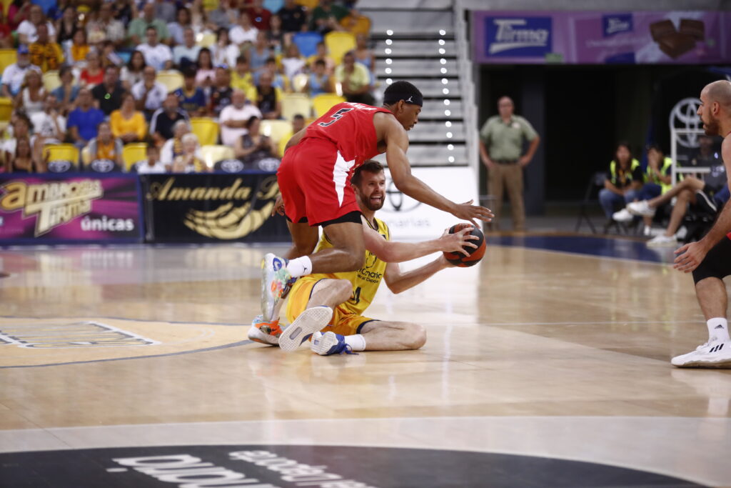 Jugadores en duelo por el balón durante un partido de baloncesto, con público de fondo.