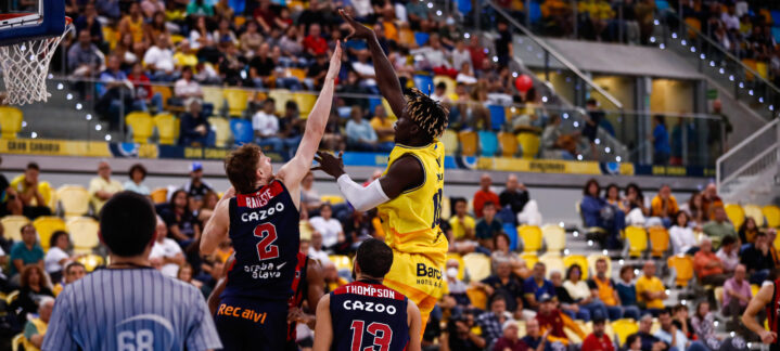 Jugador del Gran Canaria lanza a canasta mientras dos oponentes intentan bloquearlo.