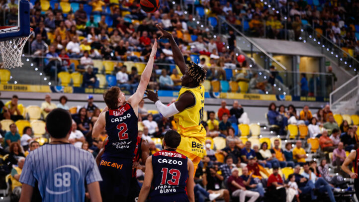 Jugador del Gran Canaria lanza a canasta mientras dos oponentes intentan bloquearlo.