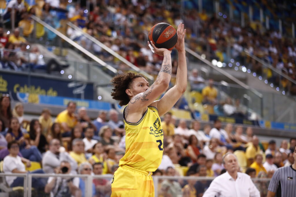Jugador del CB Gran Canaria en acción, lanzando el balón ante una multitud animada.