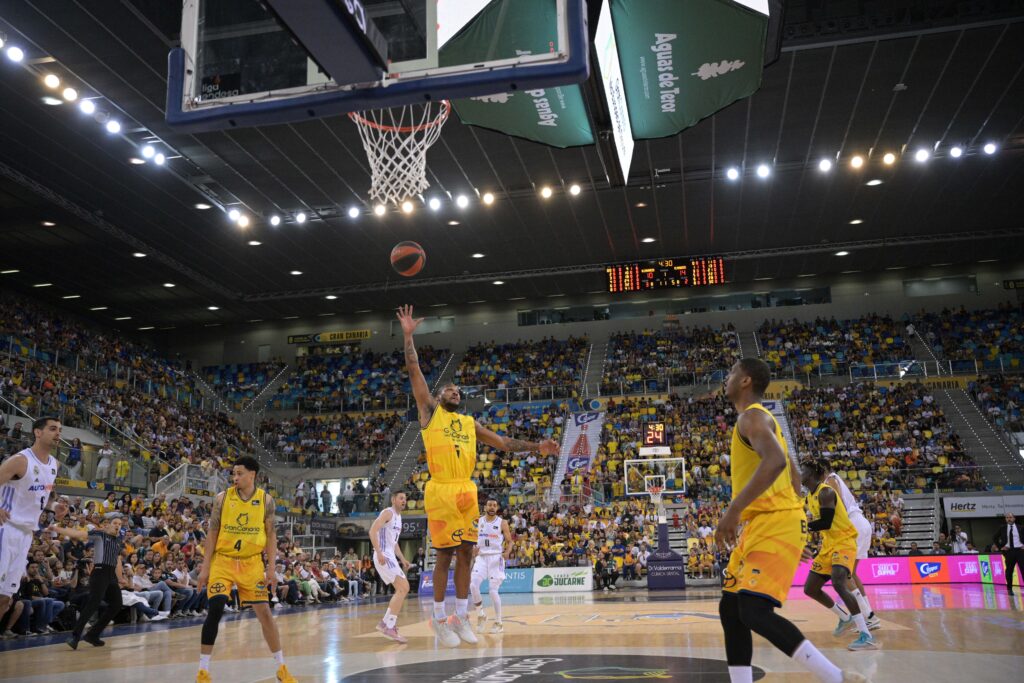 Jugador lanzando el balón hacia la canasta en un partido de baloncesto en casa.