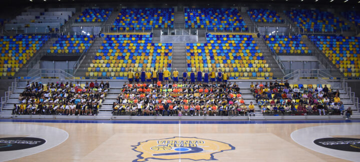 Cerca de 300 niños y niñas sentados en gradas de un pabellón de baloncesto.