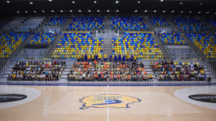 Cerca de 300 niños y niñas sentados en gradas de un pabellón de baloncesto.