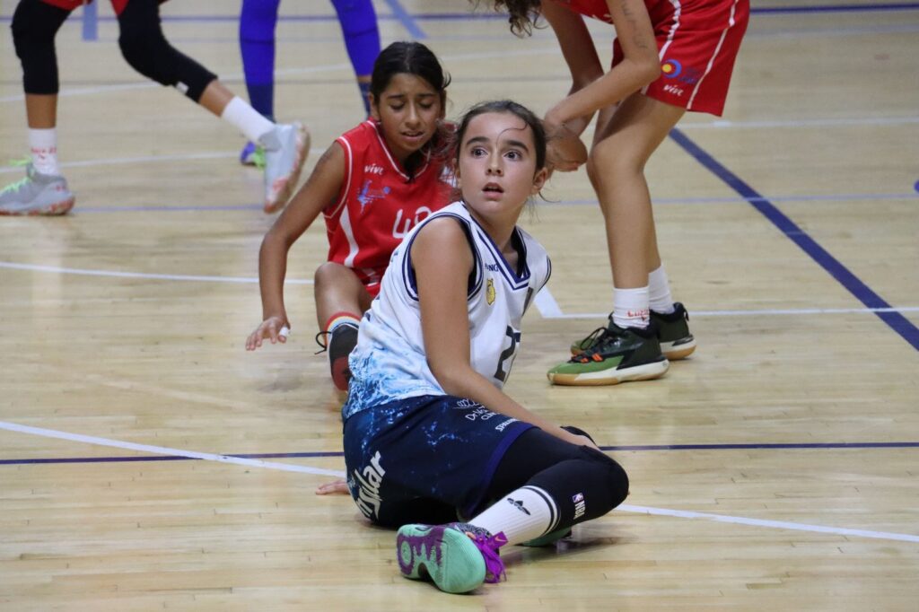 Niña sentada en la pista de baloncesto durante un partido, mirada sorpresa y compañeras alrededor.