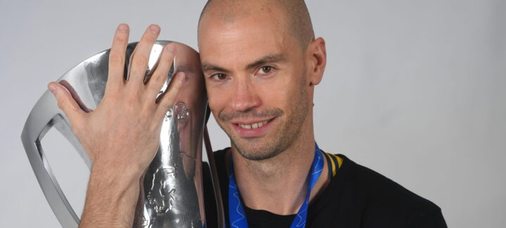 Oliver Stevic sostiene un trofeo, luciendo una medalla y sonriendo ante la cámara.