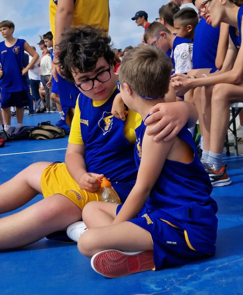 Dos niños sentados en una pista de baloncesto, conversando y compartiendo un momento cercano.