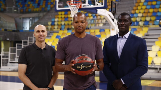 Tres hombres posan en una cancha de baloncesto, uno sostiene un balón, con fondo de gradas.