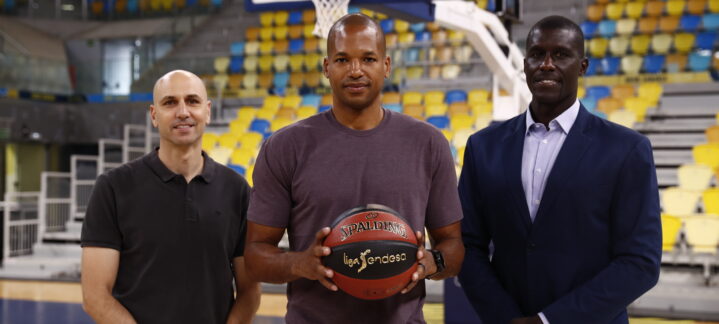 Tres hombres posan en una cancha de baloncesto, uno sostiene un balón, con fondo de gradas.