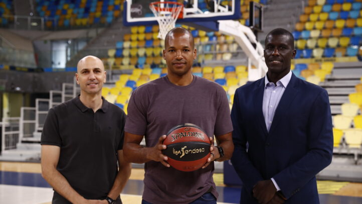 Tres hombres posan en una cancha de baloncesto, uno sostiene un balón, con fondo de gradas.
