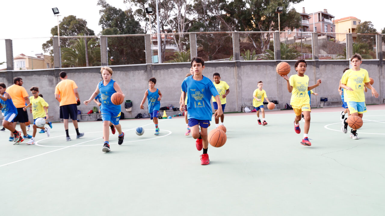 Niños entrenando baloncesto en una cancha, distintos colores de camisetas y pelotas.