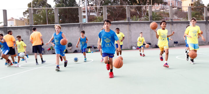 Niños entrenando baloncesto en una cancha, distintos colores de camisetas y pelotas.