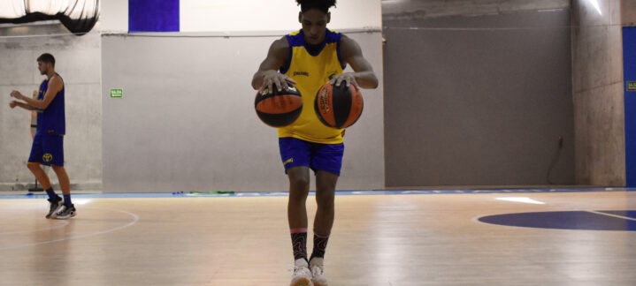 Jugador entrenando en la cancha con dos balones de baloncesto. Fondo con otros jugadores.