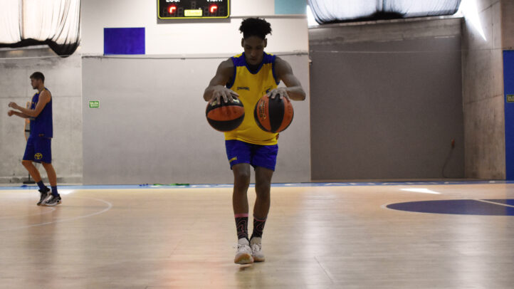 Jugador entrenando en la cancha con dos balones de baloncesto. Fondo con otros jugadores.
