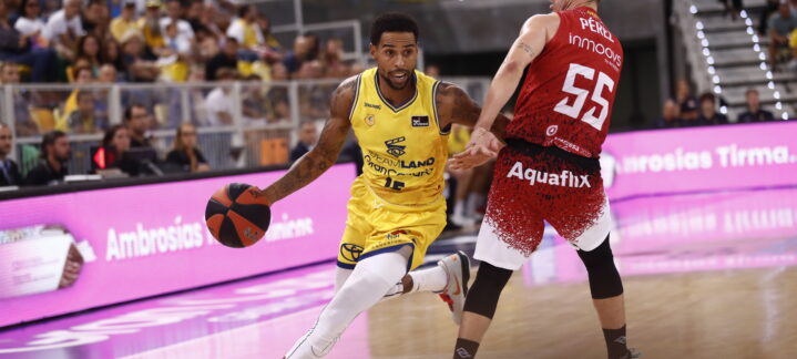 Jugador driblando el balón durante un partido de baloncesto con espectadores al fondo.