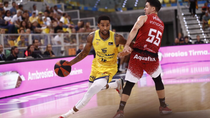 Jugador driblando el balón durante un partido de baloncesto con espectadores al fondo.
