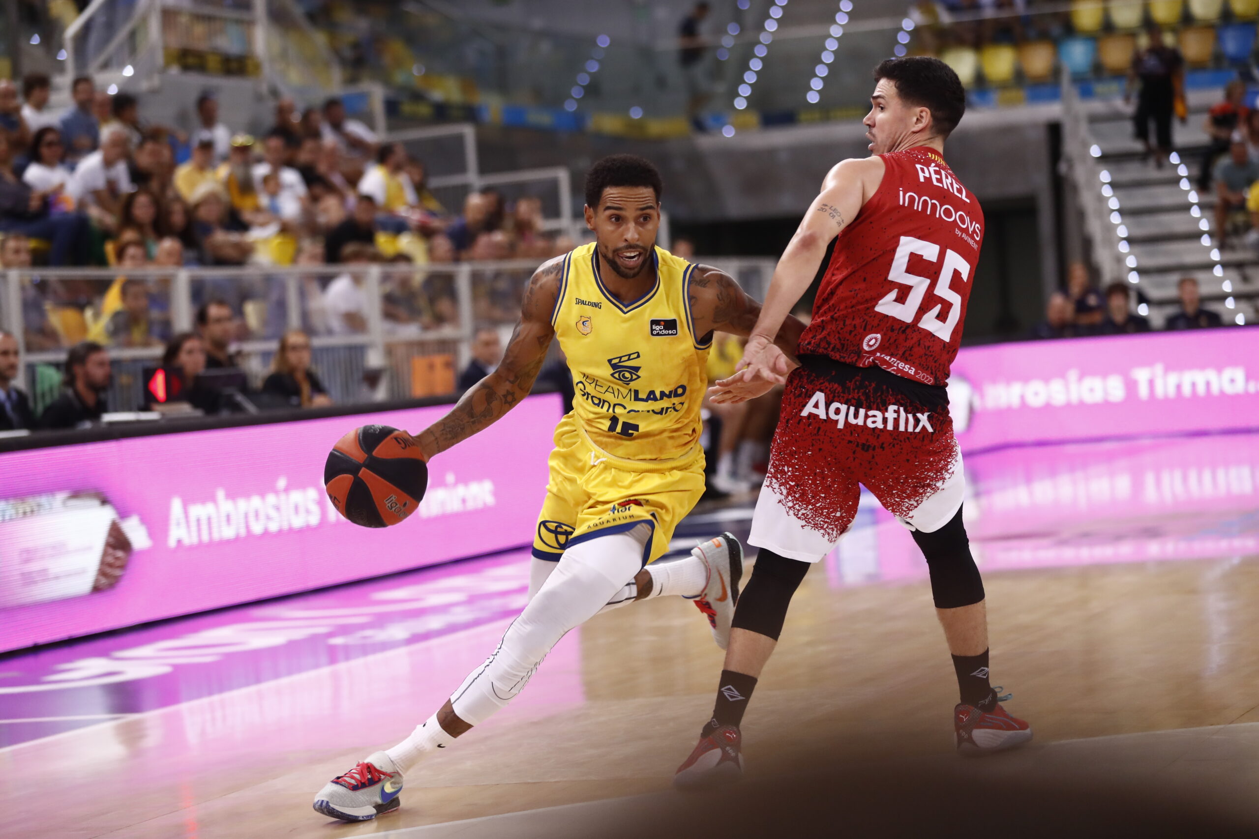 Jugador driblando el balón durante un partido de baloncesto con espectadores al fondo.