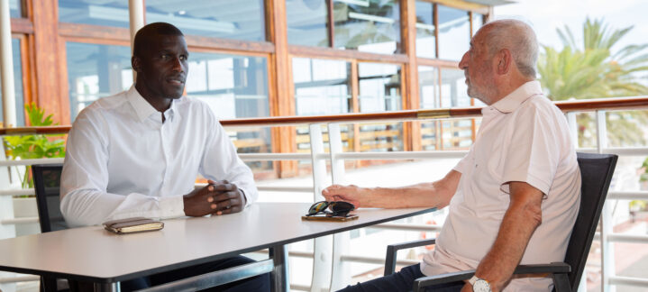 60 años de vida del Club Baloncesto Gran Canaria",Dos hombres conversan en una terraza con vista. Uno lleva camisa blanca y el otro camiseta.