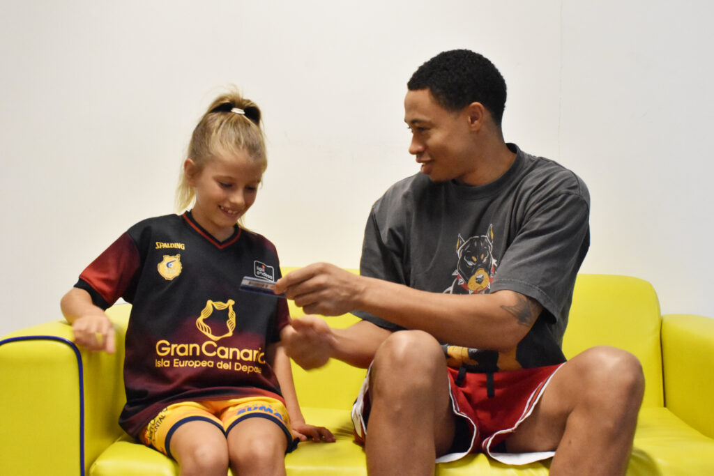Niña y jugador del Gran Canaria sonríen sentados en un sofá amarillo.