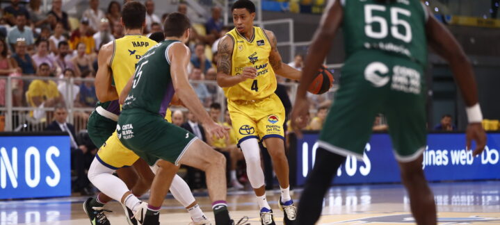 Jugador con balón en un partido de baloncesto, rodeado por defensores.