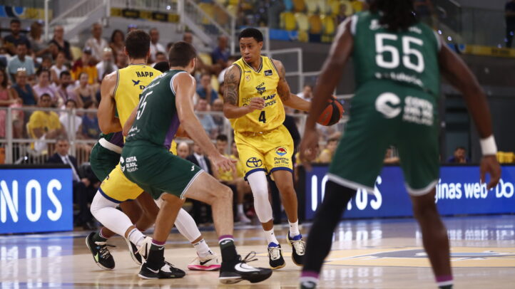Jugador con balón en un partido de baloncesto, rodeado por defensores.