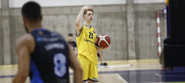 Jugadores de baloncesto en el campo, uno lanza con un balón en mano, luciendo uniforme amarillo.