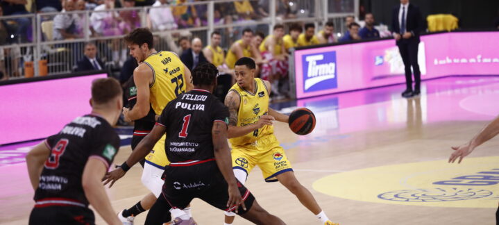 Jugadores en acción durante un partido de baloncesto, con público animando al fondo.