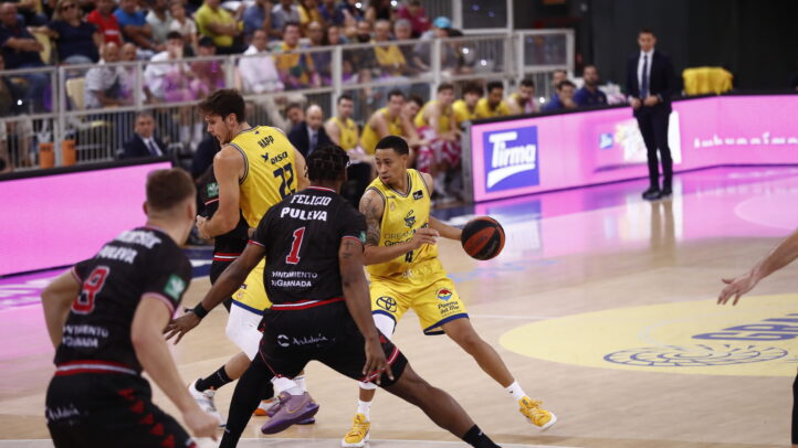 Jugadores en acción durante un partido de baloncesto, con público animando al fondo.