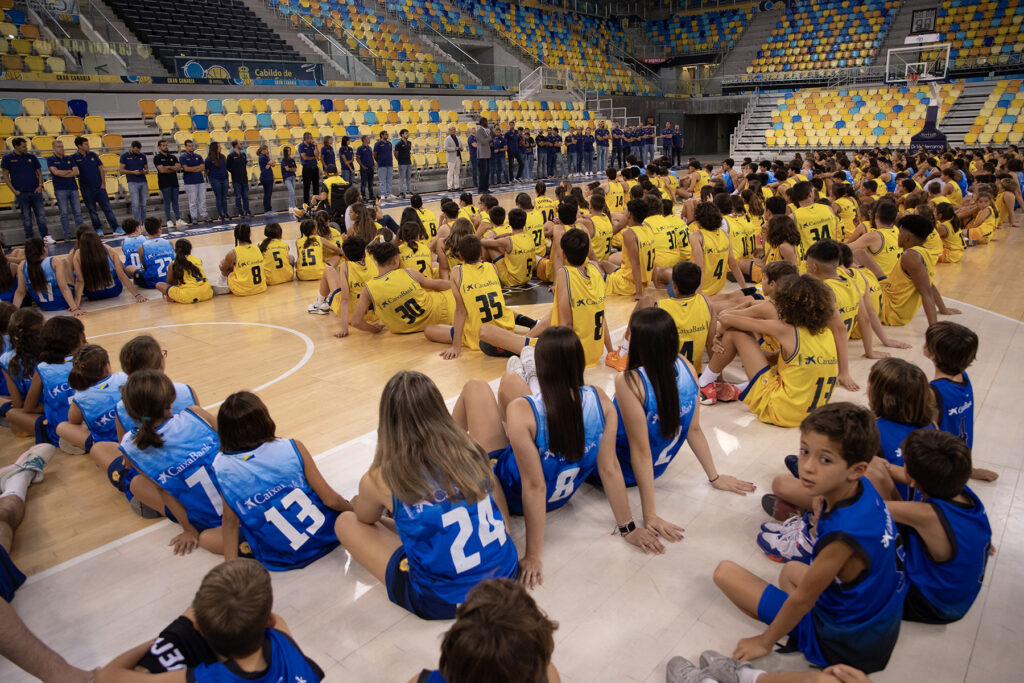 Niños y niñas sentados en el parquet, escuchando a un grupo de oradores en un evento.